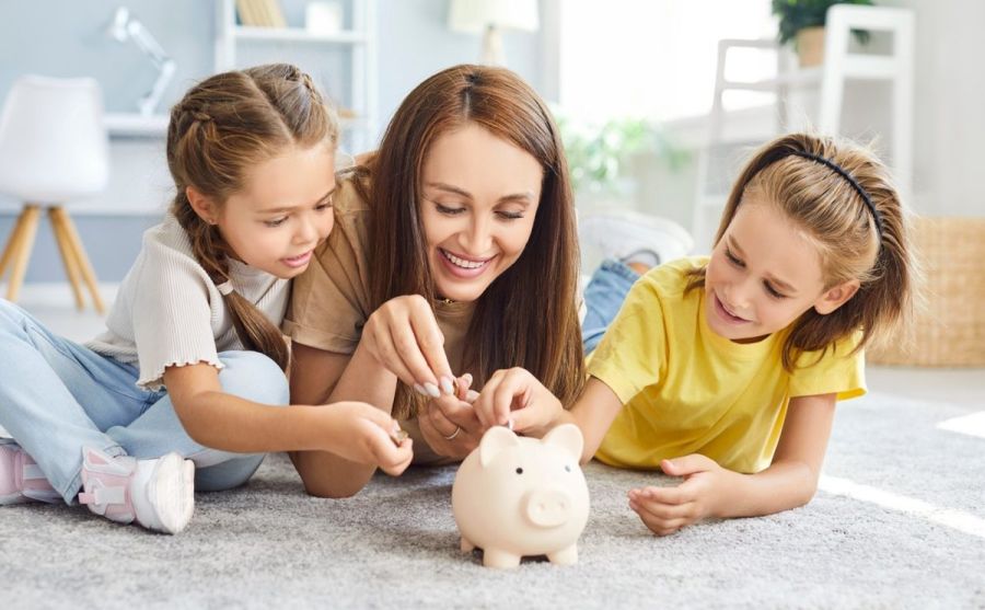 Portrait of happy family of mother and two kids putting money coin into piggy bank lying on floor in living room at home together. Family savings, investment and money management concept.